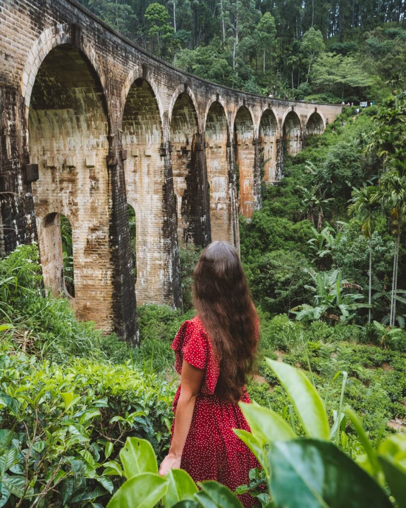 The Nine Arches Bridge in Ella, Sri Lanka, with a blue train crossing over a stone viaduct surrounded by lush green tea country.